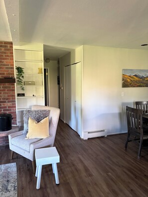 Un salón con un sillón gris, un taburete blanco, una mesa de comedor con sillas, una chimenea y una estantería.