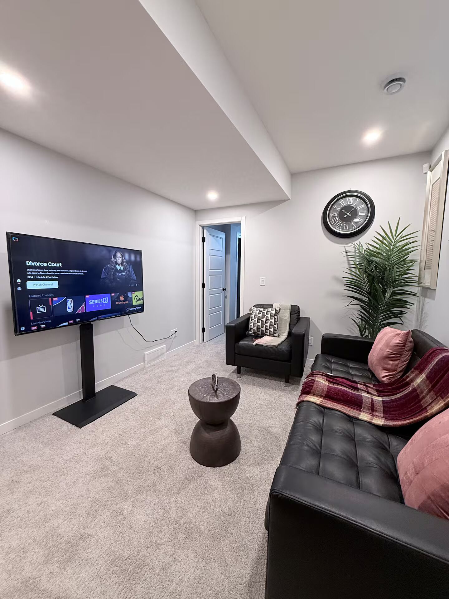 A modern living room with a flat-screen TV, a black leather sofa, a coffee table, and a potted plant.