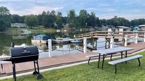 Un área verde con mesas de picnic y una parrilla, un muelle con barcos y un lago con árboles al fondo.