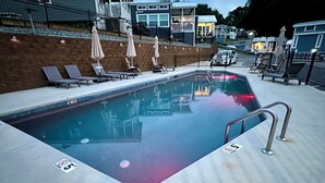Una piscina con sillas de descanso y sombrillas, un cartel de prohibición de saltos y una barandilla.