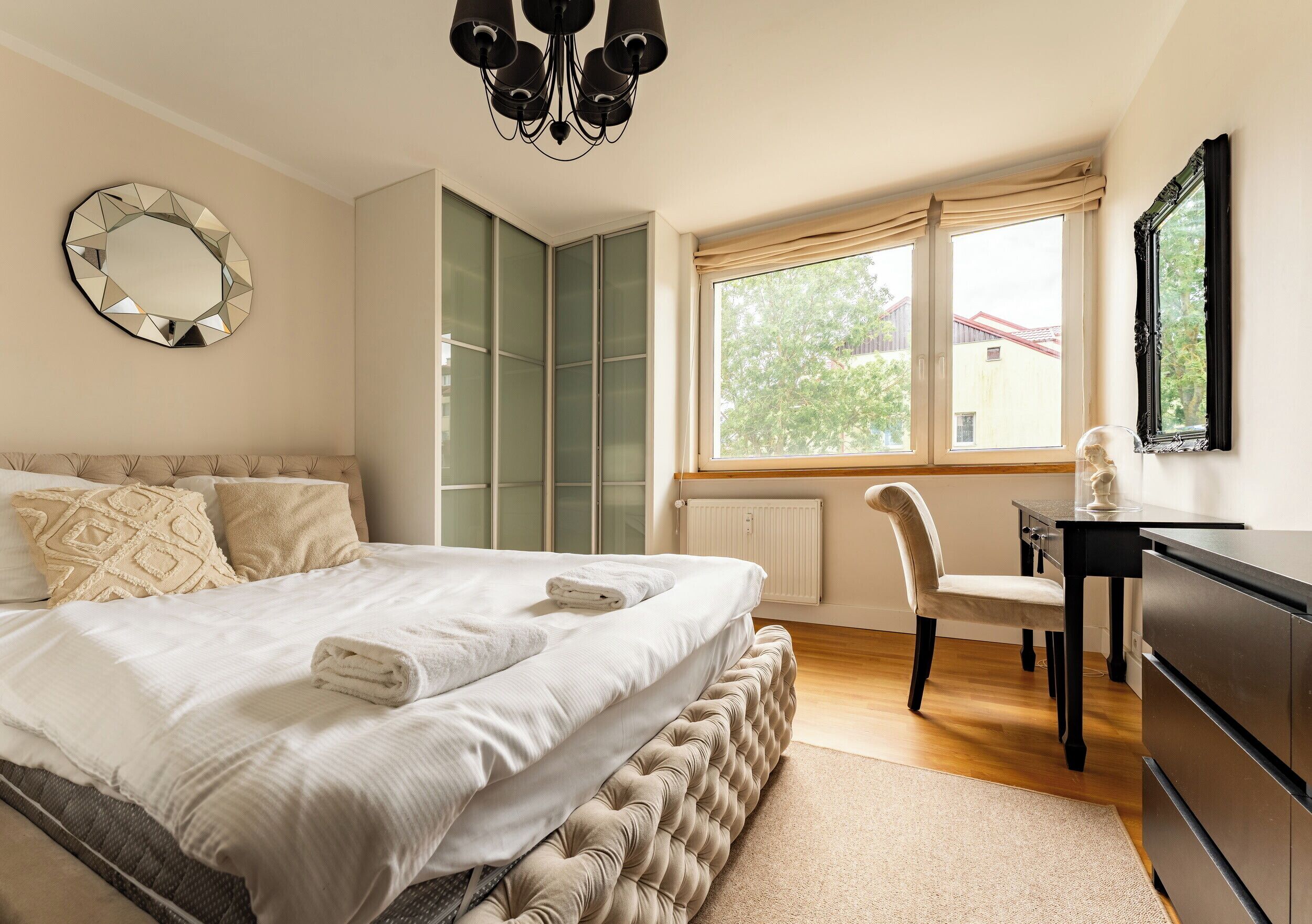 A bedroom with a bed, a desk, a chair, a dresser, a mirror, and a window with a view of trees and buildings.