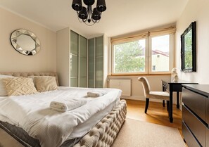 A bedroom with a bed, a desk, a chair, a dresser, a mirror, and a window with a view of trees and buildings.