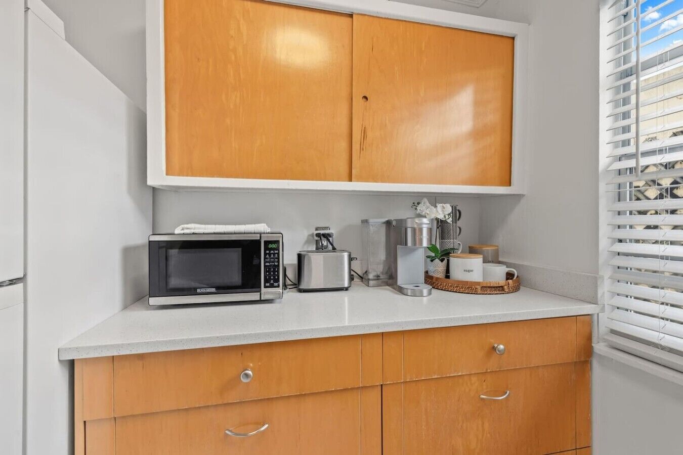 A kitchenette with a microwave, toaster, and coffee maker on a white countertop, wooden cabinets below, and a window with blinds.