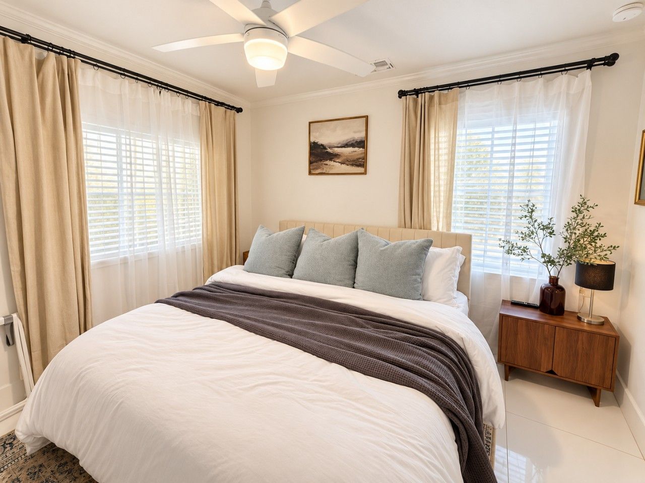 Quarto com cama, cortinas, ventilador de teto, mesa de cabeceira com abajur e janela com persianas.
