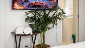 A neatly arranged room with a bed, a small table with towels, a potted plant, and a television displaying a cozy scene. - La Casita Serena | King Bed Near the Beach (West Palm Beach)