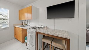 A compact kitchen with wooden cabinets, a white stove, a marble countertop, and a flat-screen TV mounted on the wall. - La Casita Serena | King Bed Near the Beach (West Palm Beach)