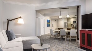 Camera d'albergo con zona living che include un divano, un tavolino e una TV. Si nota anche un'area pranzo con sedie e tavolo, e una cucina sullo sfondo.