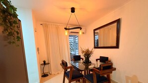 A dining area with a glass table, wooden chairs, a hanging light fixture, and a window with curtains. - Las Americas Apartment 3-bedroom with AC and Heather in Santo Domingo Este. (Santo Domingo Este)