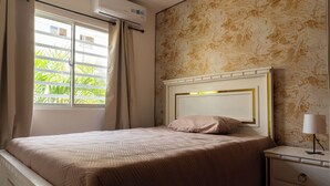 A bedroom with a bed, a nightstand, a lamp, a window with curtains, and an air conditioning unit. - Las Americas Apartment 3-bedroom with AC and Heather in Santo Domingo Este. (Santo Domingo Este)