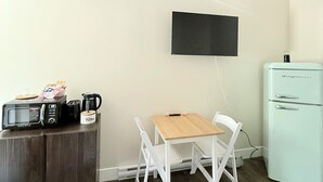 Una pequeña kitchenette con microondas, cafetera y refrigerador antiguo. Hay una mesa de madera con dos sillas blancas y un televisor de pantalla plana montado en la pared.