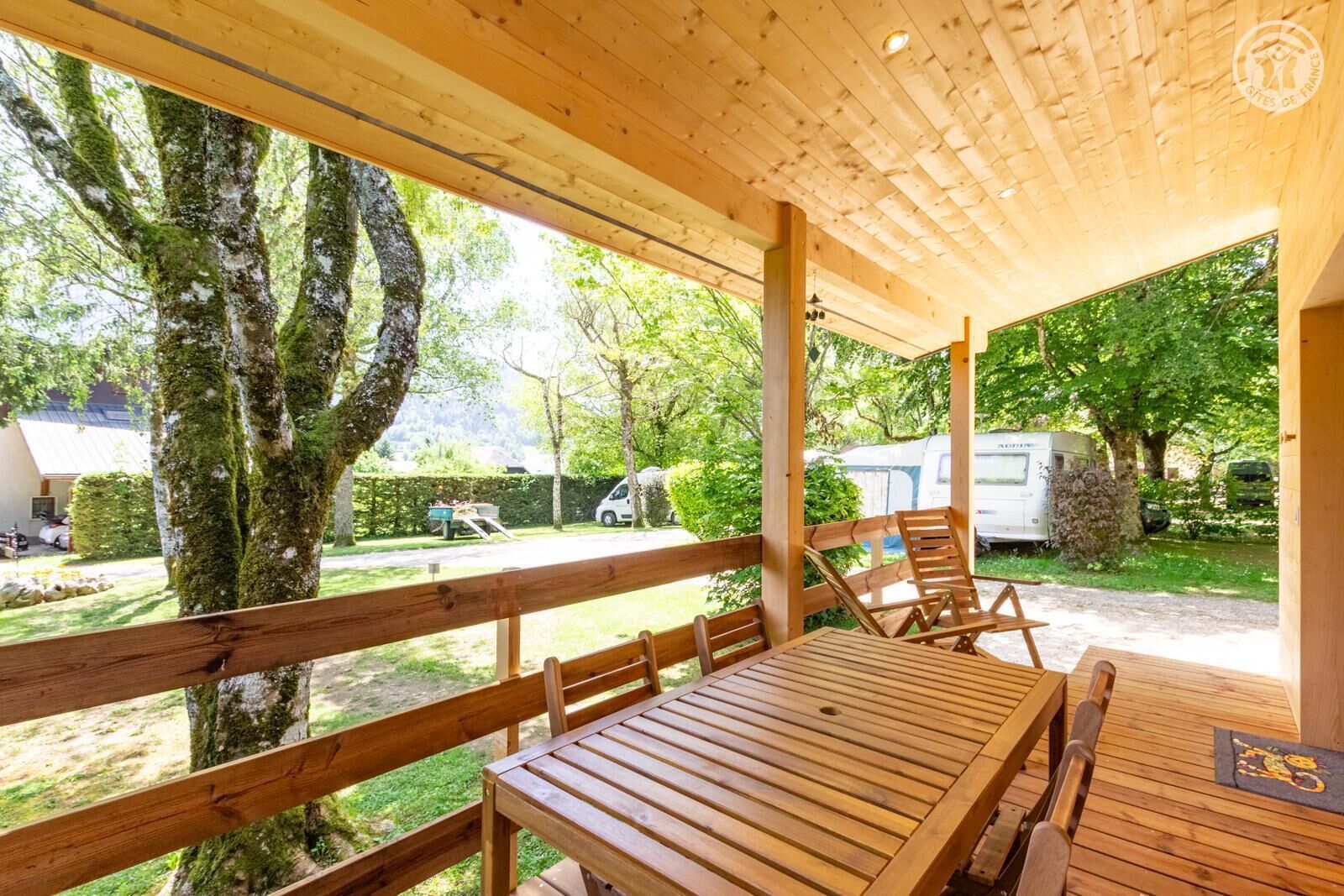 A covered outdoor seating area with wooden furniture and a view of the surrounding greenery and parked vehicles.