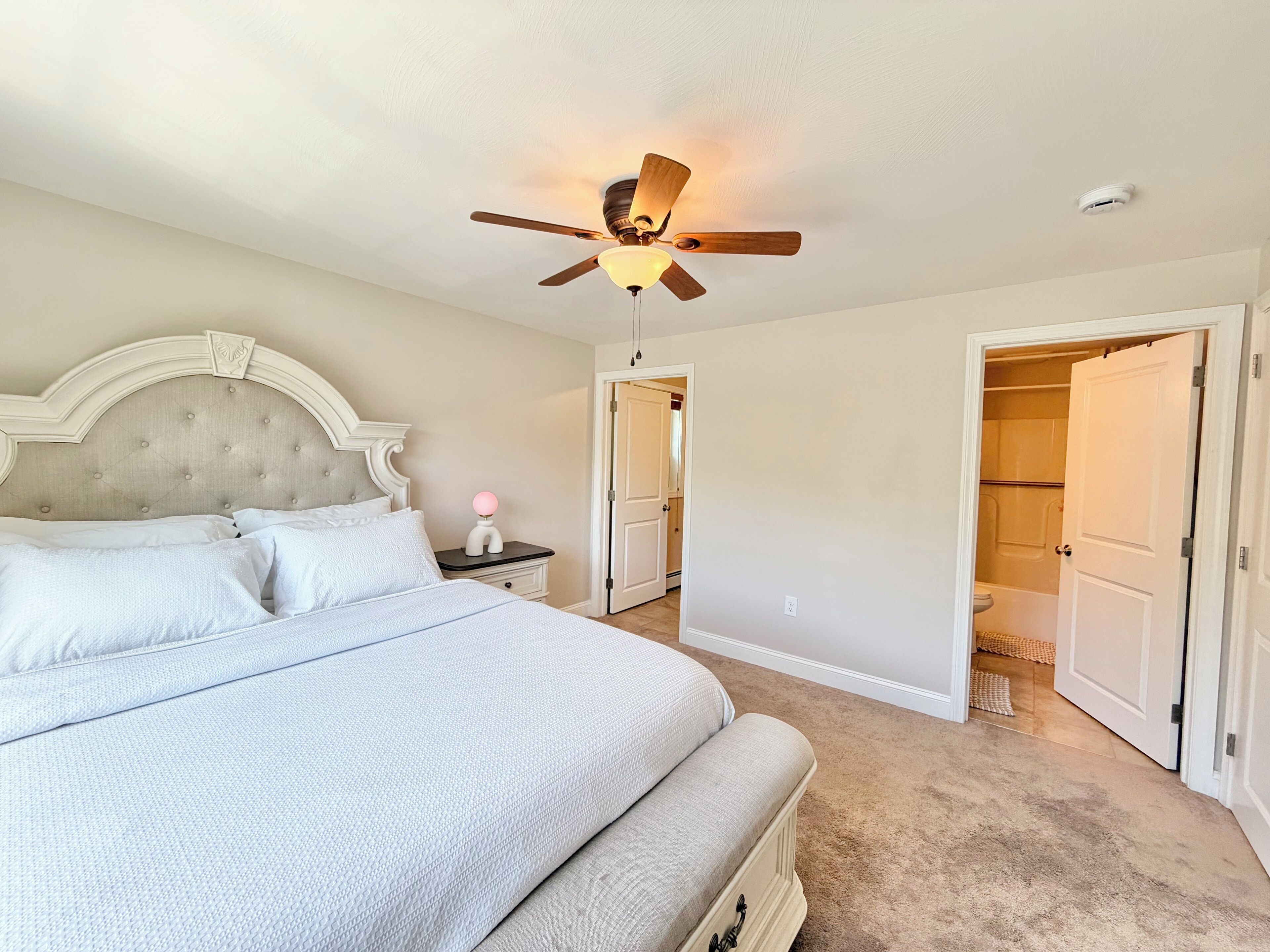 A bedroom with a bed, a nightstand, a ceiling fan, and a door leading to a bathroom.