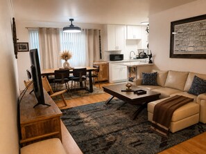 A modern living room with a sofa, coffee table, TV, and dining area with chairs and a table. - Coastal 1BR Walk to Beach Workspace 30+ Day Stays (Long Beach)