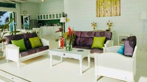 A living room with white furniture, a dining area, and a kitchen in the background.