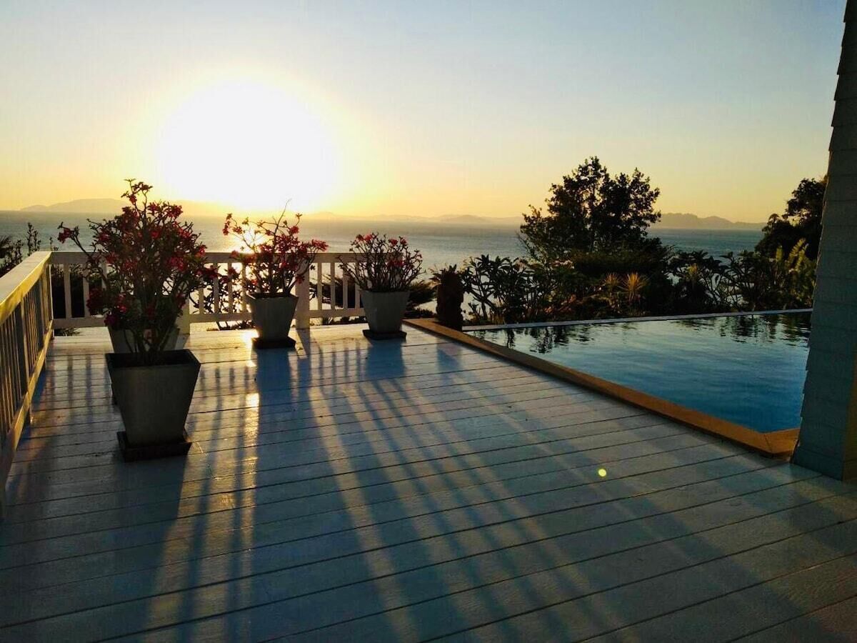 Une terrasse en bois avec une piscine, des plantes en pot et une vue sur la mer au coucher du soleil.