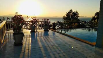 Une terrasse en bois avec une piscine, des plantes en pot et une vue sur la mer au coucher du soleil.
