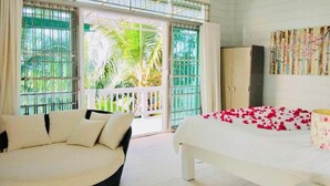 A bedroom with a bed, a sofa, and a view of a balcony with palm trees.