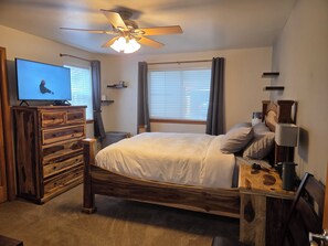 A bedroom with a wooden bed, a television on a wooden dresser, a ceiling fan, and a window with blinds. - The Royal Shreddar - Remote Work Ready (Frisco)