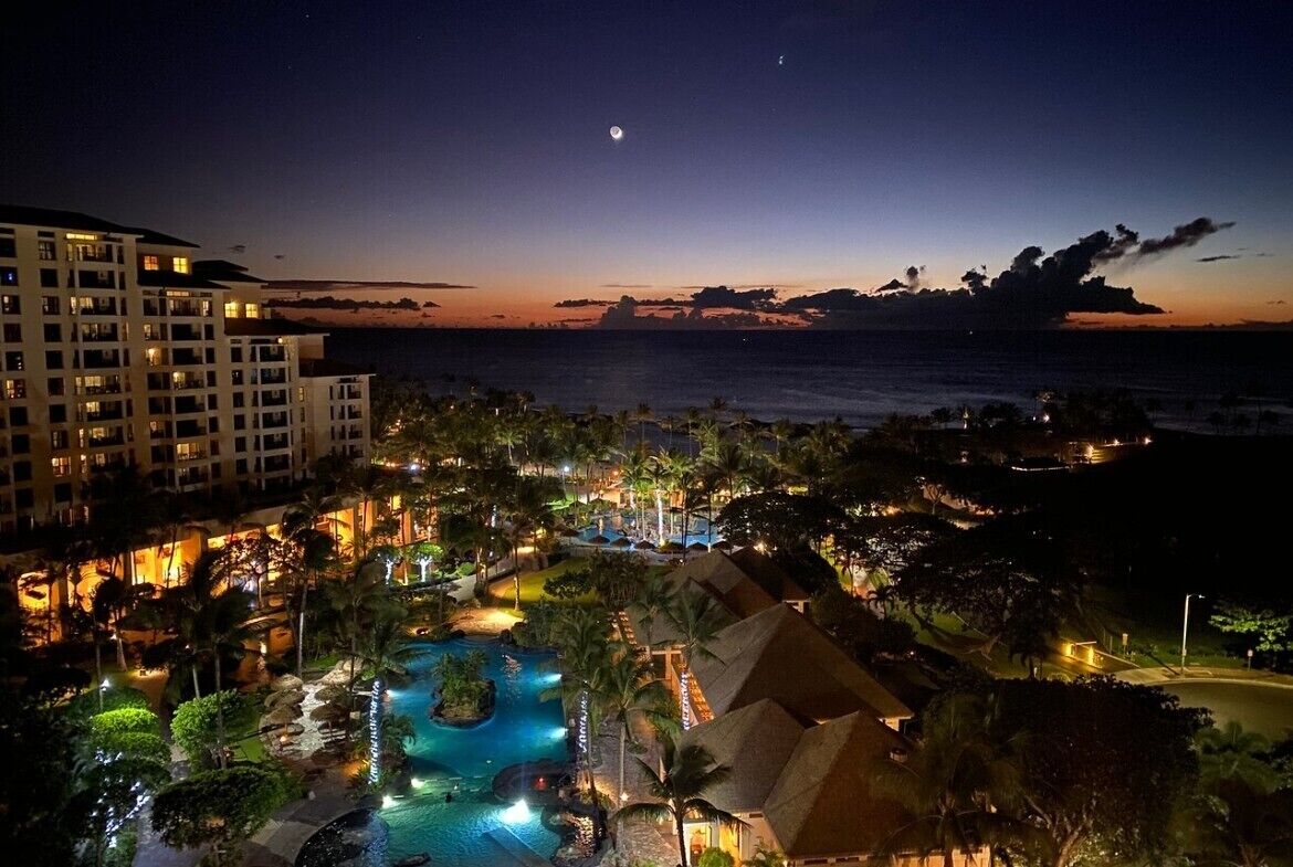 A resort with a large swimming pool, beachfront views, and a night sky with a visible moon.