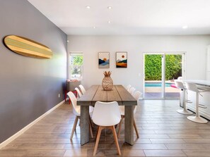 A dining area with a wooden table, white chairs, and a surfboard decoration on the wall. - Cool Pool/Spa Retreat near Shops & Surf Club (Palm Springs)