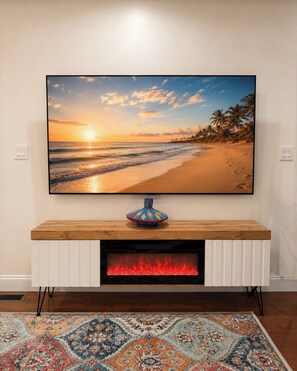 A flat-screen TV mounted on a wall above a wooden console table with a decorative vase, a fireplace below, and a patterned rug on the floor. - Cozy Room Near JFK, Train, Beach & St. John’s Hospital (Far Rockaway)