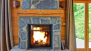 Une cheminée en pierre avec un feu de bois, des murs en bois et une vue sur la verdure à l’extérieur.
