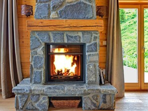 A stone fireplace with a fire, wooden walls, and a view of greenery outside. - Perle des Collons by Interhome (Thyon-Les Collons)