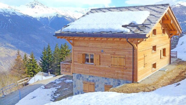 Un chalet en bois, avec des fondations en pierre, se dressant sur un fond de montagnes enneigées et d’un ciel dégagé.