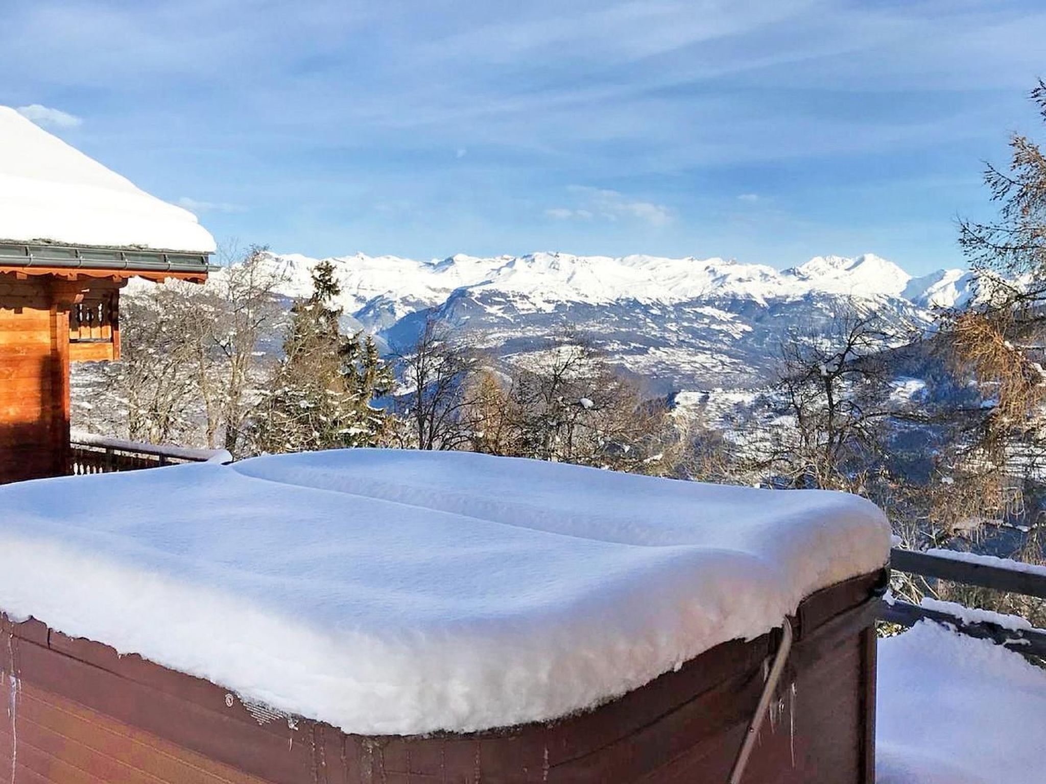 Uma cabana de madeira com telhado coberto de neve e varanda com vista para uma paisagem montanhosa nevada.