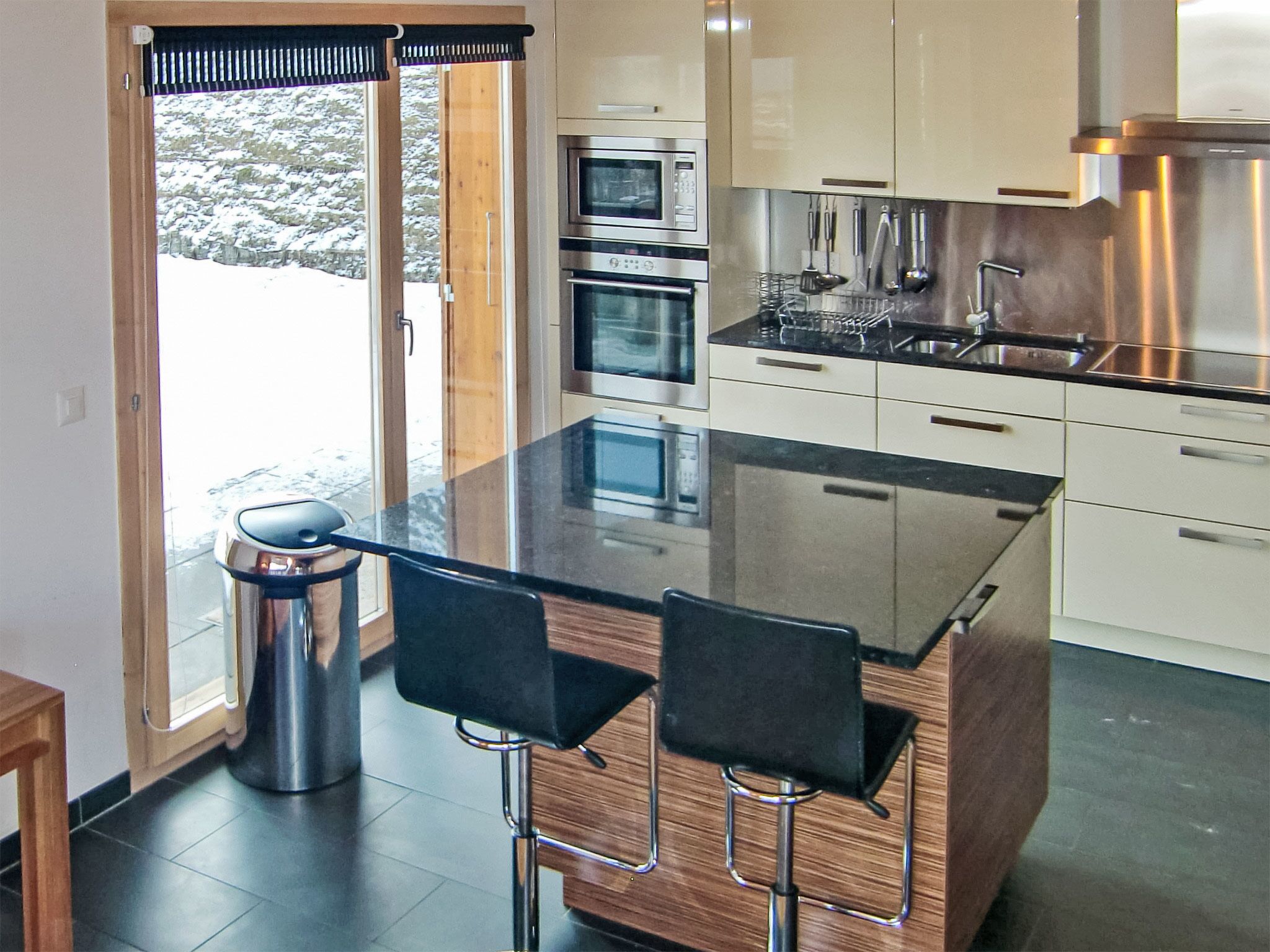 A modern kitchen with a central island, two bar stools, stainless steel appliances, and a view of a snowy landscape.