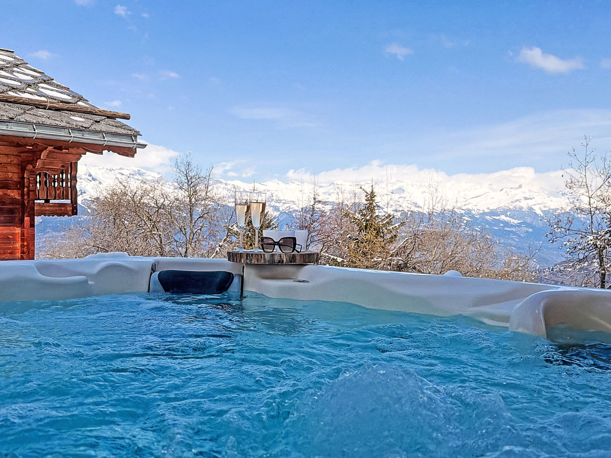 Un bain à remous avec une terrasse en bois, des verres et des lunettes de soleil, entouré de montagnes enneigées et sous un ciel dégagé.