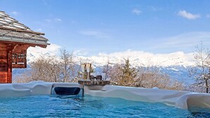 Ein Whirlpool mit Holzterrasse, Gläsern und Sonnenbrille, umgeben von schneebedeckten Bergen und blauem Himmel.