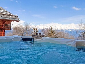 Ein Whirlpool mit Holzterrasse, Gläsern und Sonnenbrille, umgeben von schneebedeckten Bergen und blauem Himmel.