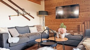 Ein modernes Wohnzimmer mit grauem Sofa, einer Holzwand, einem Flachbildfernseher und einem kleinen runden Tisch mit Blumen.