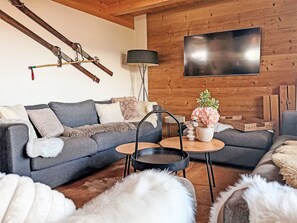 Ein modernes Wohnzimmer mit grauem Sofa, einer Holzwand, einem Flachbildfernseher und einem kleinen runden Tisch mit Blumen.