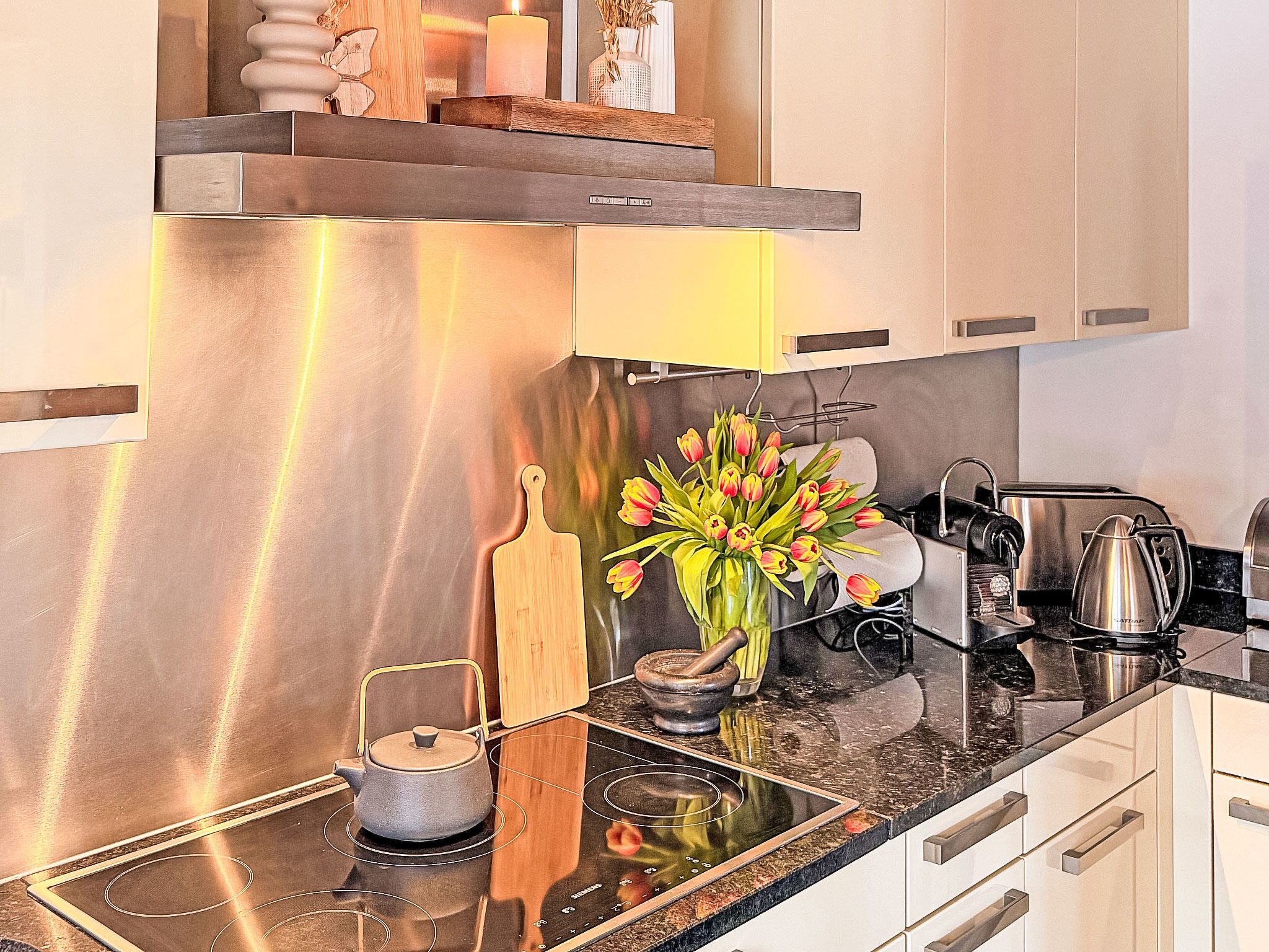 A modern kitchen with stainless steel appliances, a wooden cutting board, a mortar and pestle, a kettle, and a vase with tulips.