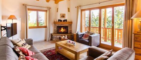 A cozy living room with a fireplace, large windows, and a wooden ceiling.
