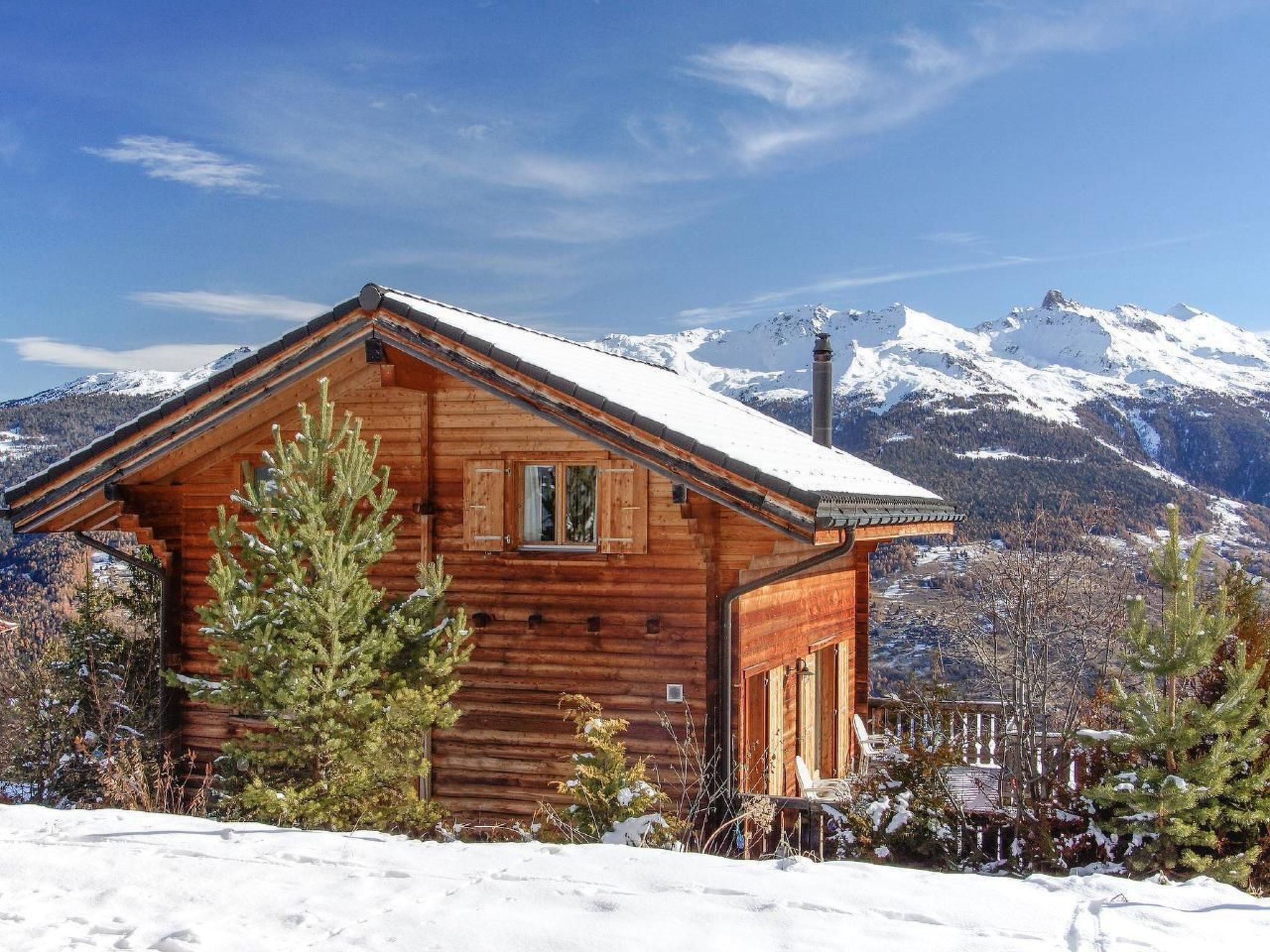 A wooden cabin with a sloped roof, surrounded by snow and trees, with a mountain range in the background.