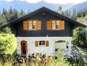 Eine Holzhütte mit dunklem Dach, zwei Fenstern mit Holzläden, einer Haustür und einem Steinweg, der dorthin führt.