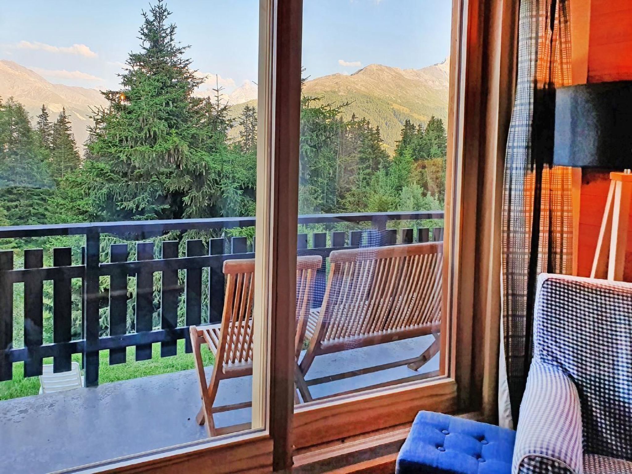 A balcony with two wooden chairs and a blue ottoman, overlooking a forested area with mountains in the background.