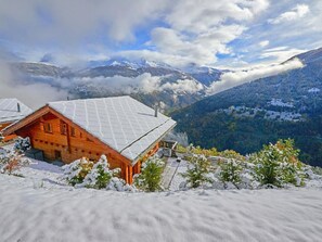 Eine Holzdiele mit schneebedecktem Dach, umgeben von verschneiter Landschaft und Bergen im Hintergrund.