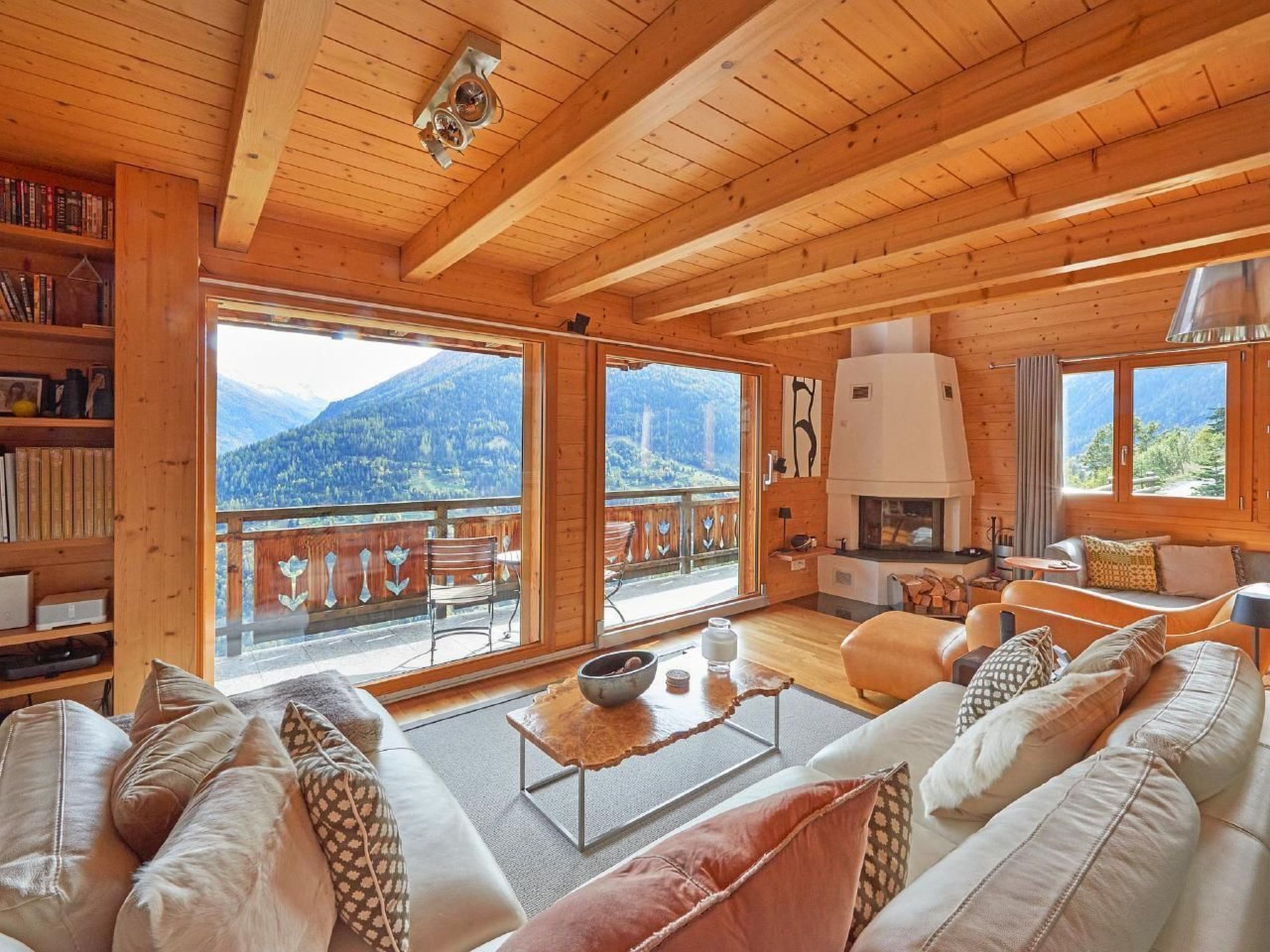 A cozy wooden cabin interior with a fireplace, large windows, and a balcony overlooking a mountainous landscape.
