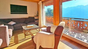 Una sala de estar acogedora con sofá, sillón y mesa de centro, un balcón con vistas a montañas, y vigas de madera en el techo.
