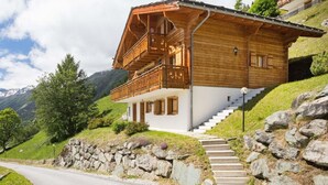 Una cabaña de madera con balcón, escalones de piedra y montañas al fondo.