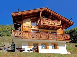 A two-story wooden chalet with balconies, a dining area with outdoor seating, and a stone wall in the background. - Etoile des 4 Vallees by Interhome (Hérémence)