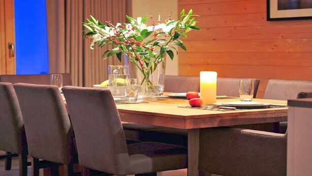 A dining area with a wooden table, chairs, a vase with flowers, a candle, and a glass of orange juice. - Chalet Falcons Nest by Interhome (Hérémence)