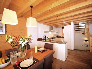 A modern kitchen with wooden beams, a dining area with a table set for two, and a staircase leading to an upper level. - Chalet Falcons Nest by Interhome (Hérémence)