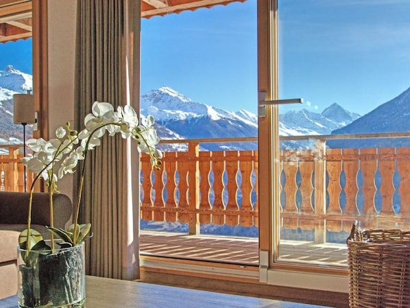 Una habitación con vistas a montañas nevadas, un balcón de madera y una mesa de vidrio con un jarrón de flores blancas.