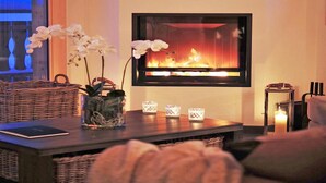 A cozy living room with a fireplace, a wicker sofa, a coffee table with decorative items, and a view of the outdoors. - Chalet Falcons Nest by Interhome (Hérémence)
