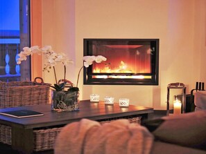 A cozy living room with a fireplace, a wicker sofa, a coffee table with decorative items, and a view of the outdoors. - Chalet Falcons Nest by Interhome (Hérémence)